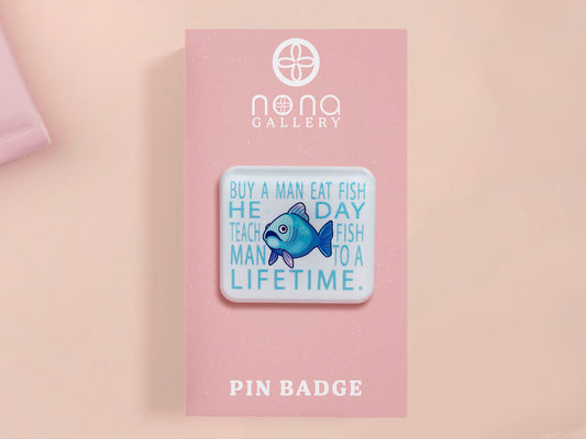 A clear acrylic pin badge with a pink heart-shaped pin back clasp. The design of the pin badge is of a cute chibi blue fish with the quote buy a man eat fish he day teach fish man to a lifetime.