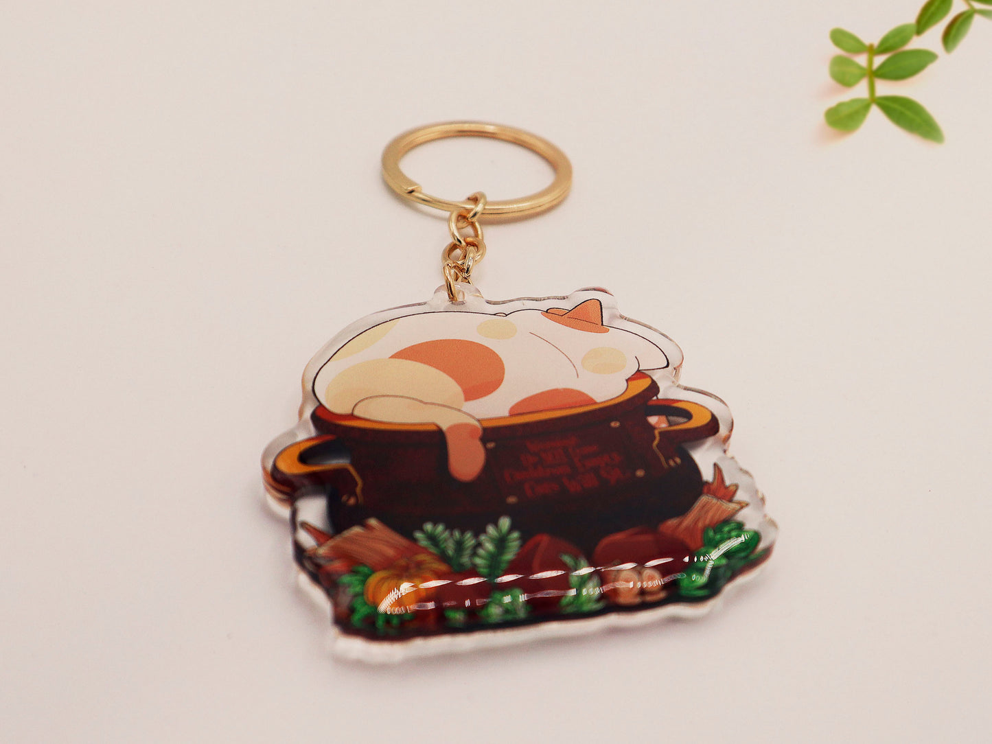 Double sided epoxy clear acrylic keychain with golden clasp of a fat calico cat sat happily inside a witches cauldron surrounded by plants and pumpkins.