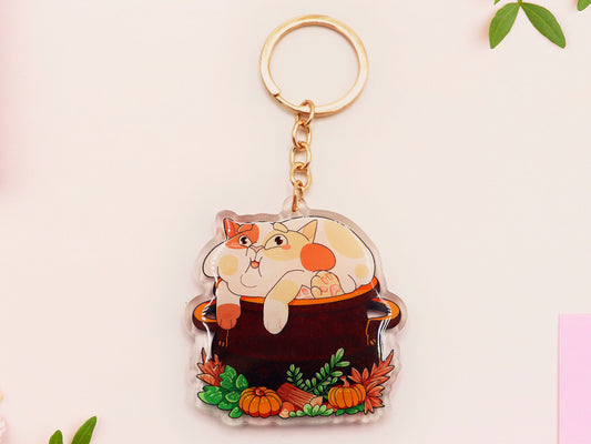 Double sided epoxy clear acrylic keychain with golden clasp of a fat calico cat sat happily inside a witches cauldron surrounded by plants and pumpkins.