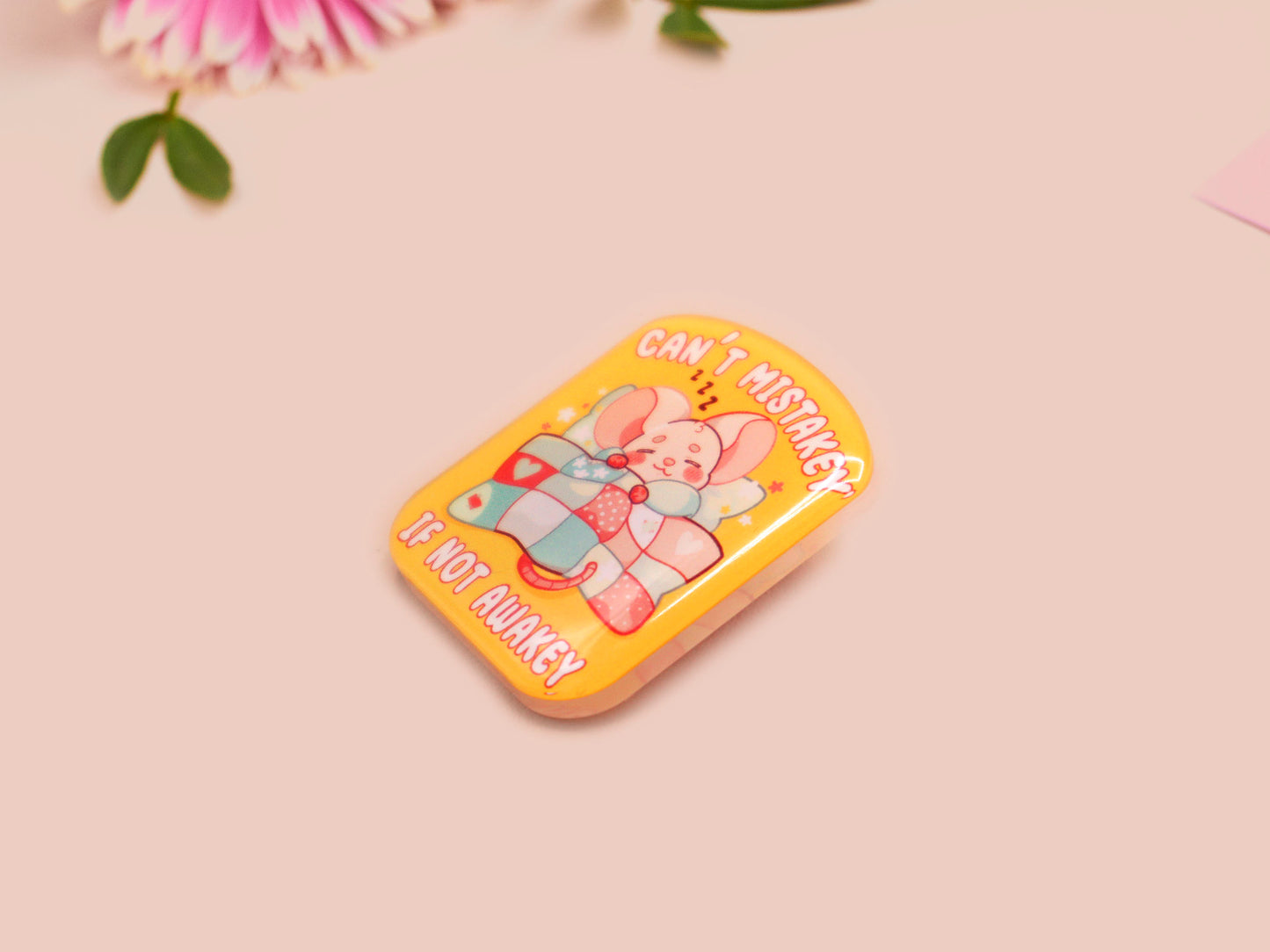 A yellow acrylic pin badge with a pink heart-shaped pin back clasp. The design of the pin badge is of a cute mouse napping in bed with a patchwork quilt with the quote can't mistakey if not awakey.