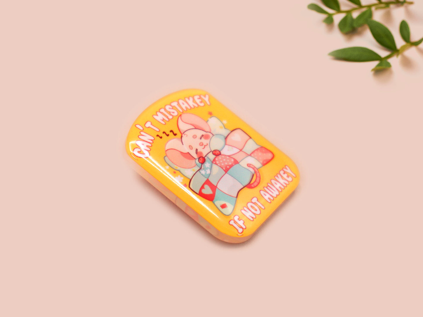 A yellow acrylic pin badge with a pink heart-shaped pin back clasp. The design of the pin badge is of a cute mouse napping in bed with a patchwork quilt with the quote can't mistakey if not awakey.