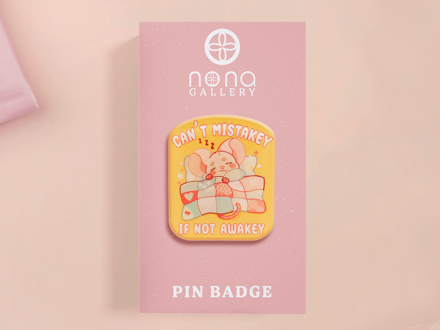 A yellow acrylic pin badge with a pink heart-shaped pin back clasp. The design of the pin badge is of a cute mouse napping in bed with a patchwork quilt with the quote can't mistakey if not awakey.