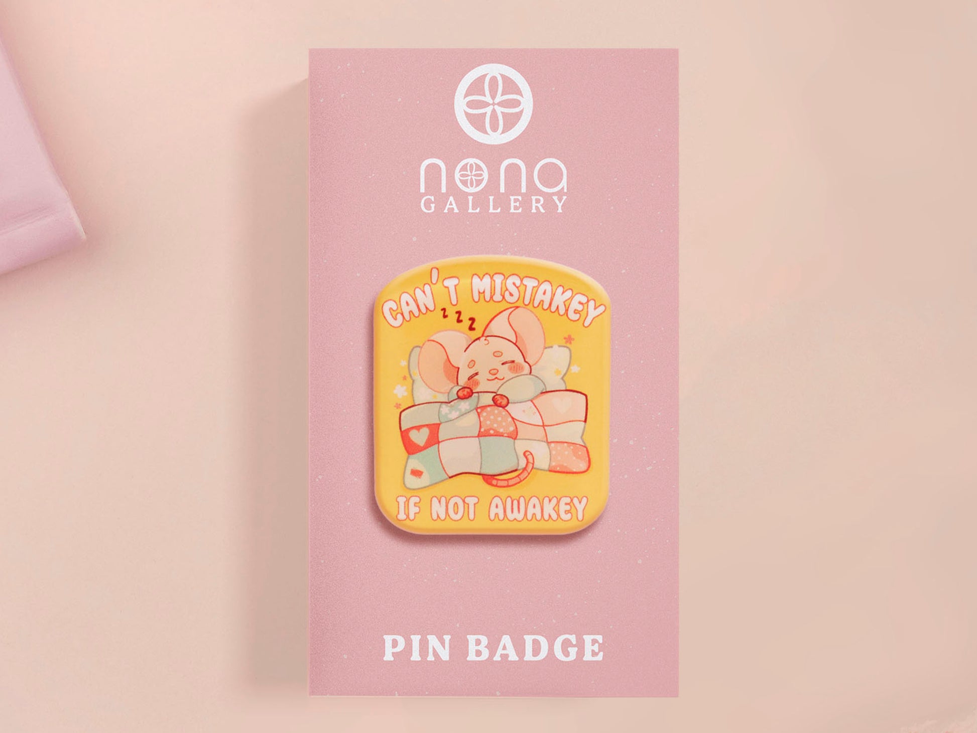 A yellow acrylic pin badge with a pink heart-shaped pin back clasp. The design of the pin badge is of a cute mouse napping in bed with a patchwork quilt with the quote can't mistakey if not awakey.