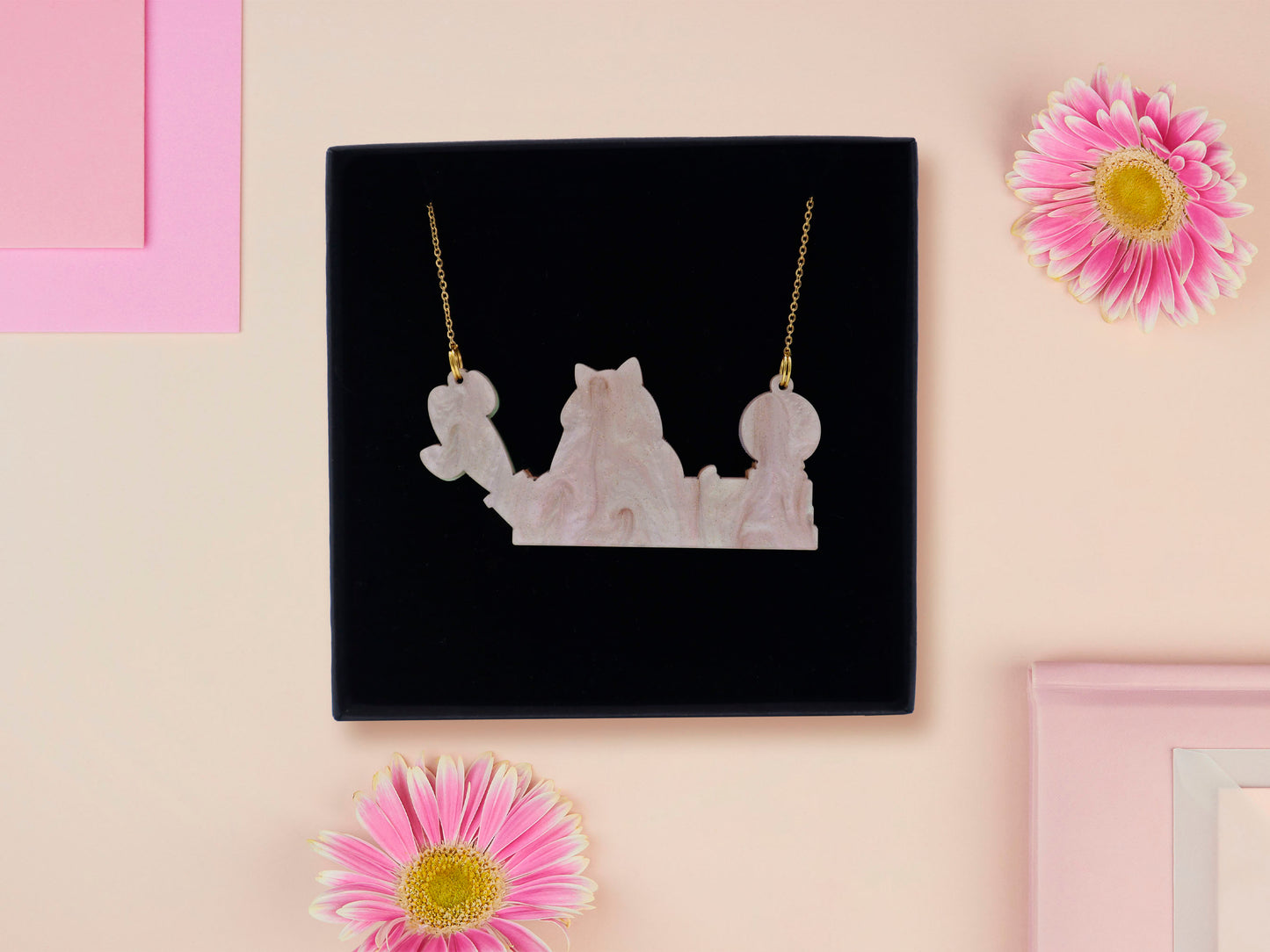 A wooden and glitter acrylic necklace with gold chain in a black box of a cute fat ginger cat sat on a shelf mischievously pushing a cactus of the end of the shelf, whilst sat next to a crystal ball and potions