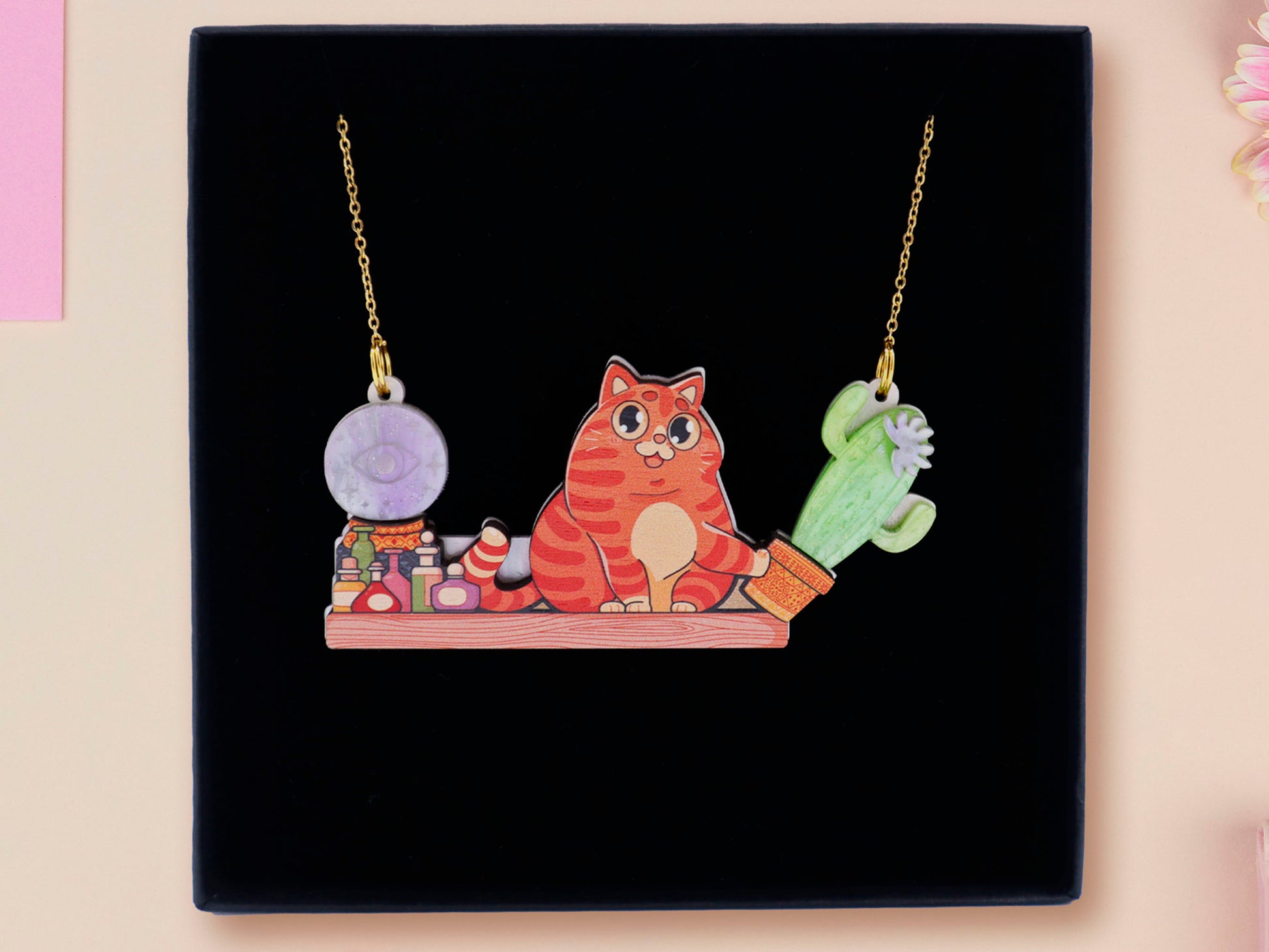 A wooden and glitter acrylic necklace with gold chain in a black box of a cute fat ginger cat sat on a shelf mischievously pushing a cactus of the end of the shelf, whilst sat next to a crystal ball and potions