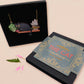 Mixed material handmade necklace of chibi cartoon cat sat on a witch's book shelf with pearlescent crystal. knocking a plant pot of the edge, with a gold chain and black gift box with a blue familiars collection gift sleeve.