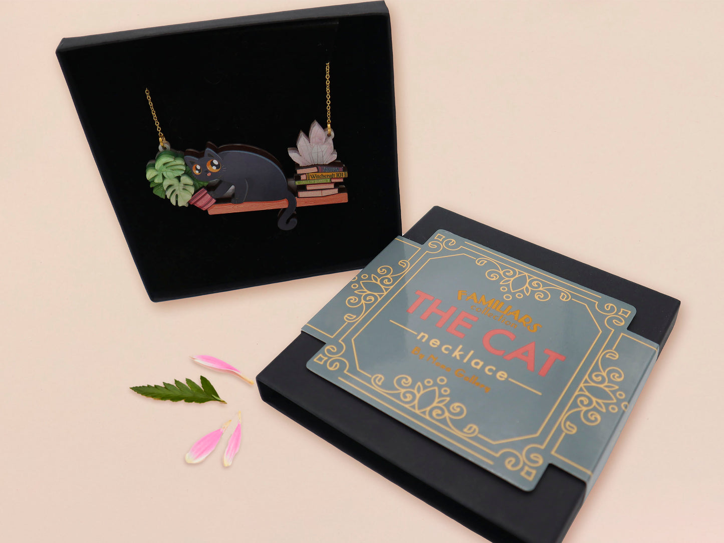 Mixed material handmade necklace of chibi cartoon cat sat on a witch's book shelf with pearlescent crystal. knocking a plant pot of the edge, with a gold chain and black gift box with a blue familiars collection gift sleeve.