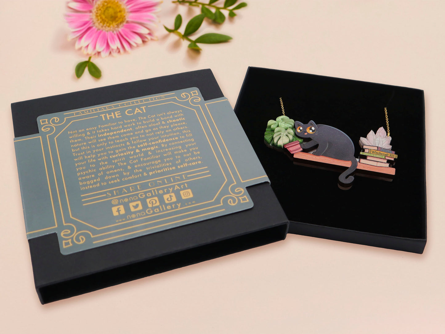 Mixed material handmade necklace of chibi cartoon cat sat on a witch's book shelf with pearlescent crystal. knocking a plant pot of the edge, with a gold chain and black gift box with a blue familiars collection gift sleeve.