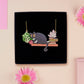 Mixed material handmade necklace of chibi cartoon cat sat on a witch's book shelf with pearlescent crystal. knocking a plant pot of the edge, with a gold chain and black gift box.
