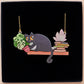 Mixed material handmade necklace of chibi cartoon cat sat on a witch's book shelf with pearlescent crystal. knocking a plant pot of the edge, with a gold chain and black gift box.