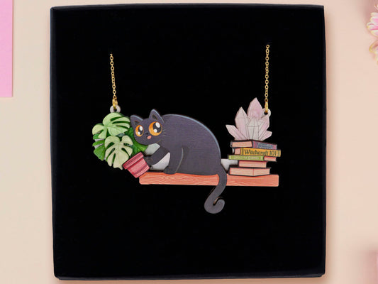 Mixed material handmade necklace of chibi cartoon cat sat on a witch's book shelf with pearlescent crystal. knocking a plant pot of the edge, with a gold chain and black gift box.