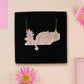 Back of mixed material handmade necklace of chibi cartoon cat sat on a witch's book shelf with pearlescent crystal. knocking a plant pot of the edge, with a gold chain and black gift box.