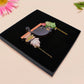 Mixed material handmade necklace of chibi cartoon cat sat on a witch's book shelf with pearlescent crystal. knocking a plant pot of the edge, with a gold chain and black gift box.