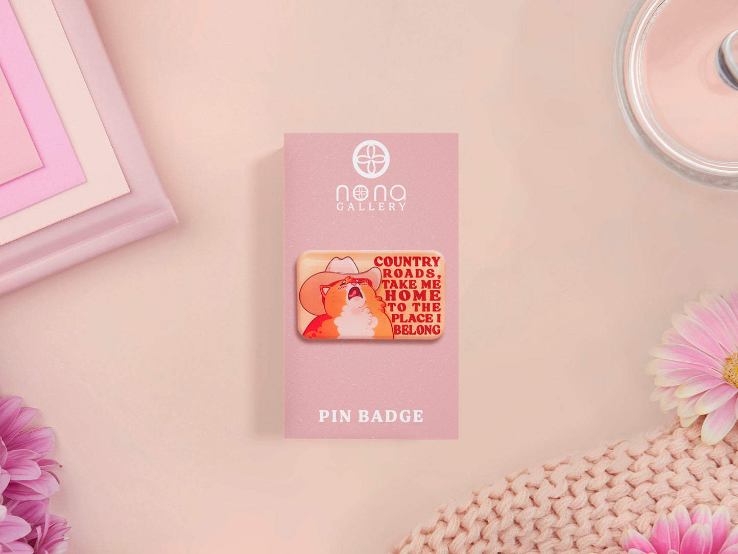 A clear acrylic pin badge with a pink heart-shaped pin back clasp. The design of the pin badge is of a cute singing cowboy cat from the viral internet meme with the song quote Country roads take me home to the place I belong