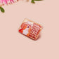 A clear acrylic pin badge with a pink heart-shaped pin back clasp. The design of the pin badge is of a cute singing cowboy cat from the viral internet meme with the song quote Country roads take me home to the place I belong