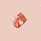 A clear acrylic pin badge with a pink heart-shaped pin back clasp. The design of the pin badge is of a cute singing cowboy cat from the viral internet meme with the song quote Country roads take me home to the place I belong