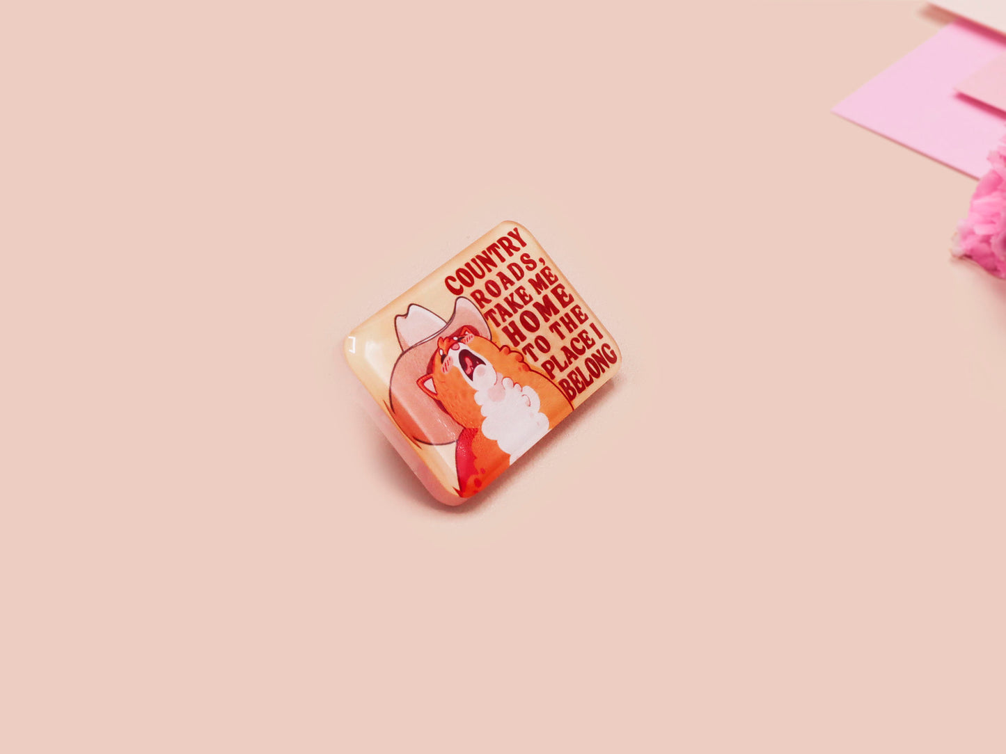 A clear acrylic pin badge with a pink heart-shaped pin back clasp. The design of the pin badge is of a cute singing cowboy cat from the viral internet meme with the song quote Country roads take me home to the place I belong
