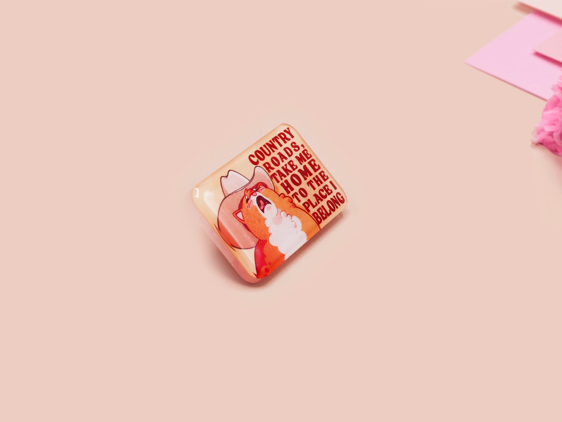 A clear acrylic pin badge with a pink heart-shaped pin back clasp. The design of the pin badge is of a cute singing cowboy cat from the viral internet meme with the song quote Country roads take me home to the place I belong