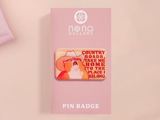 A clear acrylic pin badge with a pink heart-shaped pin back clasp. The design of the pin badge is of a cute singing cowboy cat from the viral internet meme with the song quote Country roads take me home to the place I belong