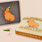 Adorable cute capybara with an orange on its head necklace made of wood and glitter pearlescent acrylic with a gold chain, displayed in a velvet gift box with a unique cute capybara sleeve design.
