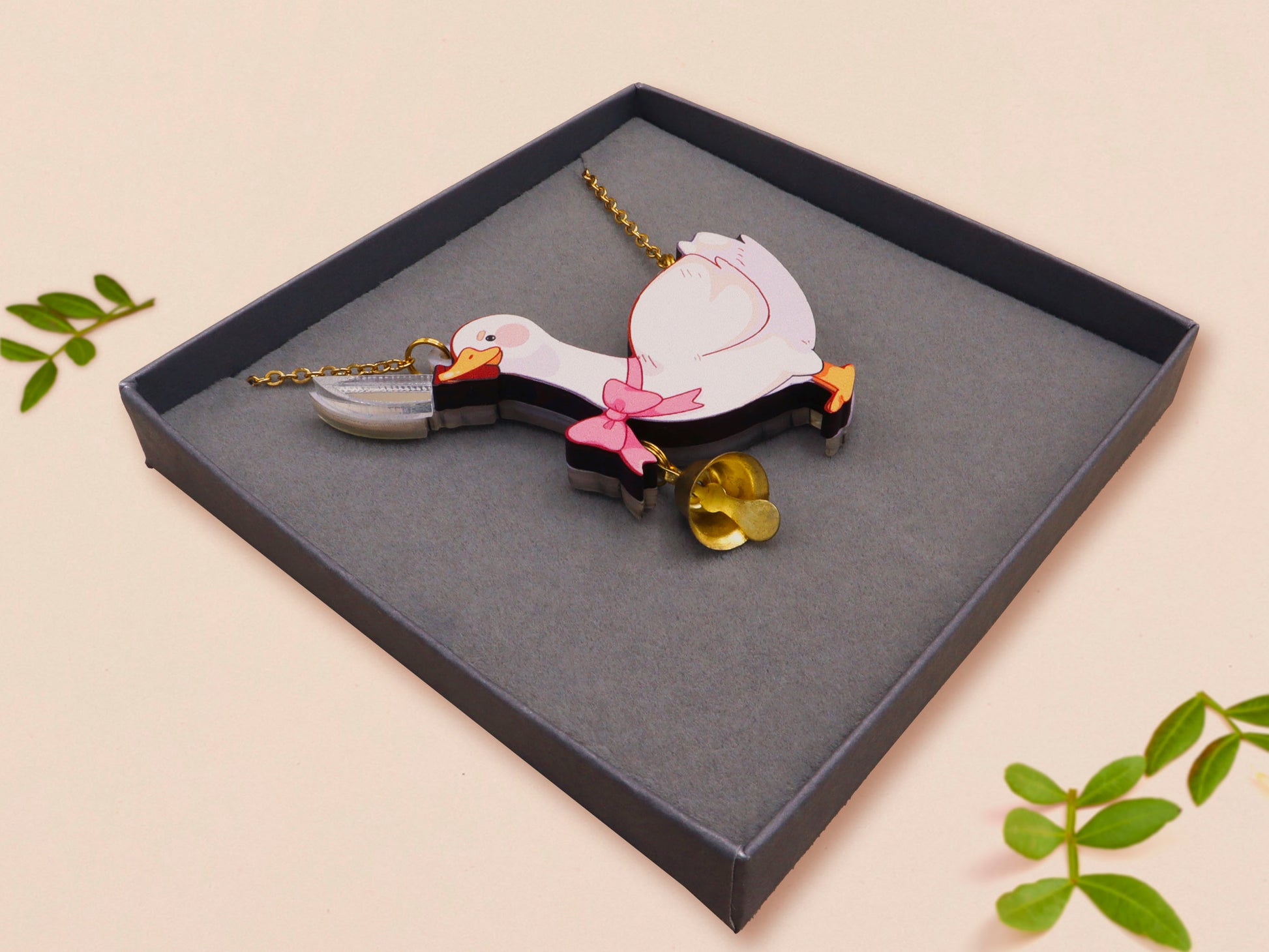 An adorable cute goose with a bell and pink bow necklace made of wood and glitter pearlescent acrylic with a gold chain is displayed in a velvet gift box with a unique cute goose sleeve design.