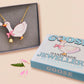 An adorable cute goose with a bell and pink bow necklace made of wood and glitter pearlescent acrylic with a gold chain is displayed in a velvet gift box with a unique cute goose sleeve design.