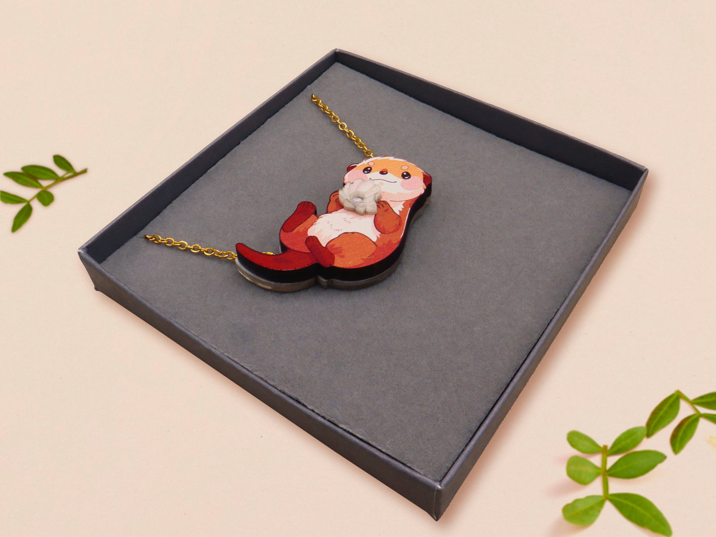 An adorable cute otter holding a clam seashell with a pearl inside necklace made of wood and glitter pearlescent acrylic with a gold chain is displayed in a velvet gift box with a unique cute otter sleeve design.