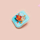 A clear acrylic pin badge with a pink heart-shaped pin back clasp. The design of the pin badge is of a cute relaxing capybara in water with a face mask with the quote don't worry be cappy.