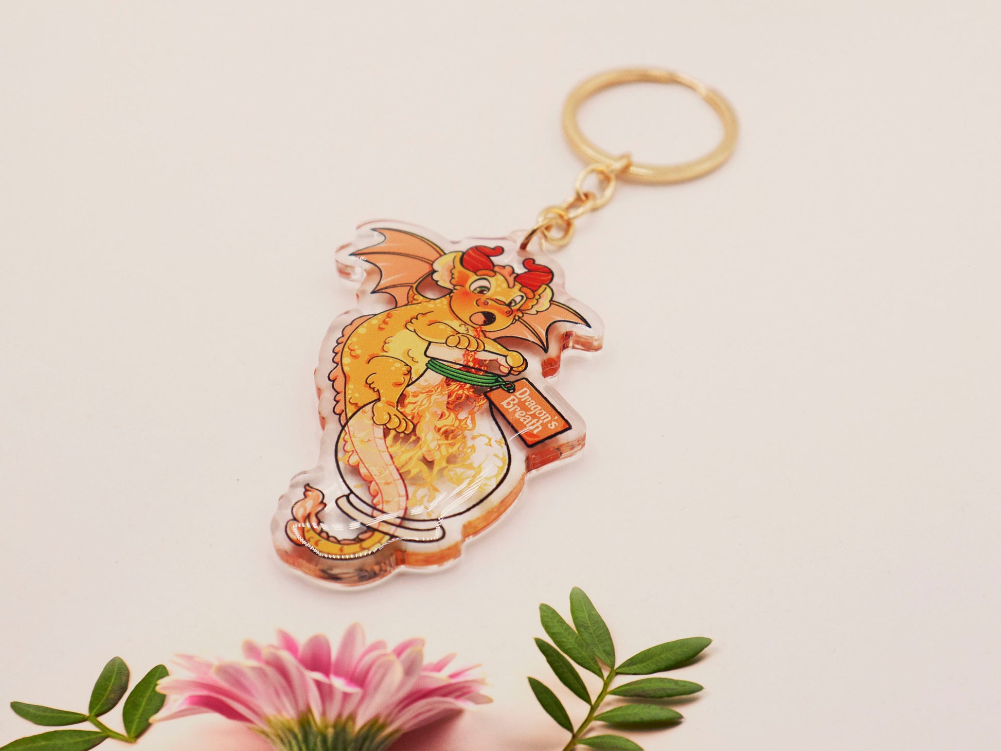 Clear acrylic keychain with gold clasp with a cartoon illustration of an orange baby dragon with a wings open hugging a potion bottle whilst breathing fire flames inside, labelled Dragon's Breath.