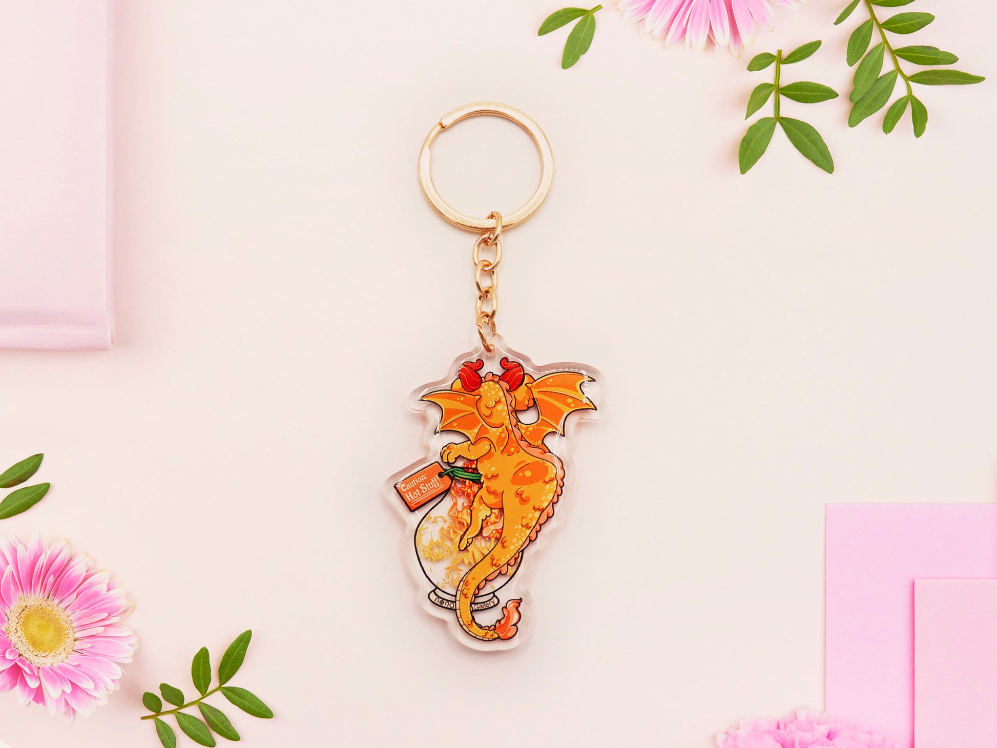 Clear acrylic keychain with gold clasp with a cartoon illustration of an orange baby dragon with a wings open hugging a potion bottle whilst breathing fire flames inside, labelled Dragon's Breath.