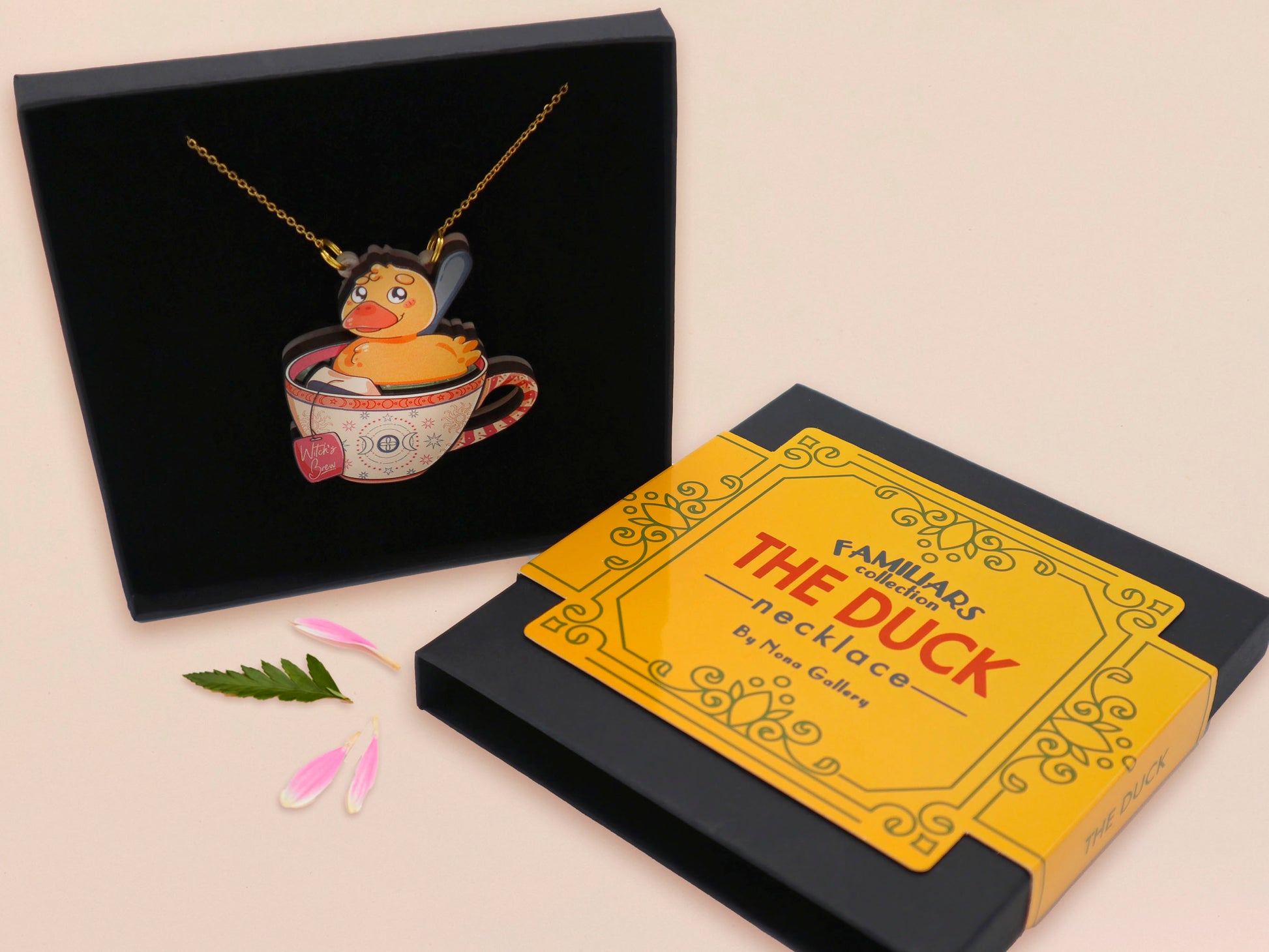 Mixed material handmade necklace of chibi cartoon duck sat in a pearlescent witch's brew teacup, with a gold chain and black gift box with a yellow familiars collection gift sleeve