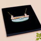 A wooden and glitter acrylic necklace with gold chain in a black box of a cute family of ducks two adult white ducks and four yellow ducklings playing and splashing in the water