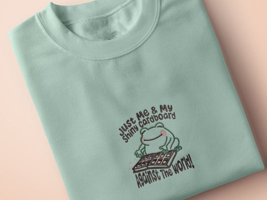 An embroidered green t-shirt design of a cute frog happily looking at his binder of tcg trading cards surrounded by the quote Just Me And My Shiny Cardboard Against The World