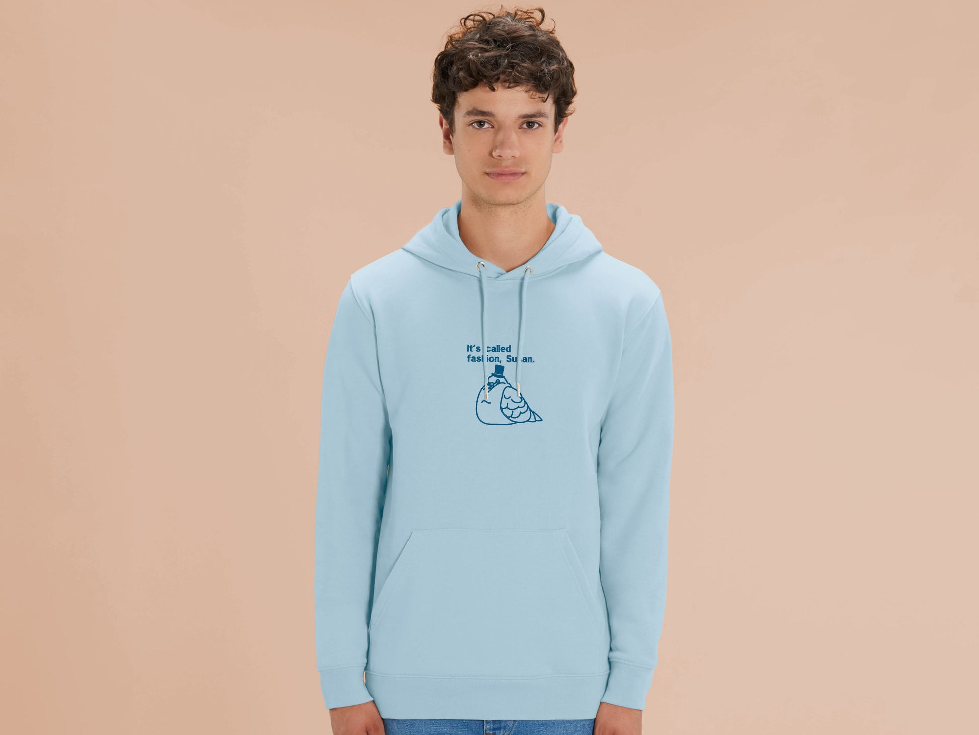 A man wearing a blue long sleeve fleece hoodie, with an embroidered blue thread design of cute fat pigeon wearing a top hat with text underneath reading It's Called Fashion, Susan.