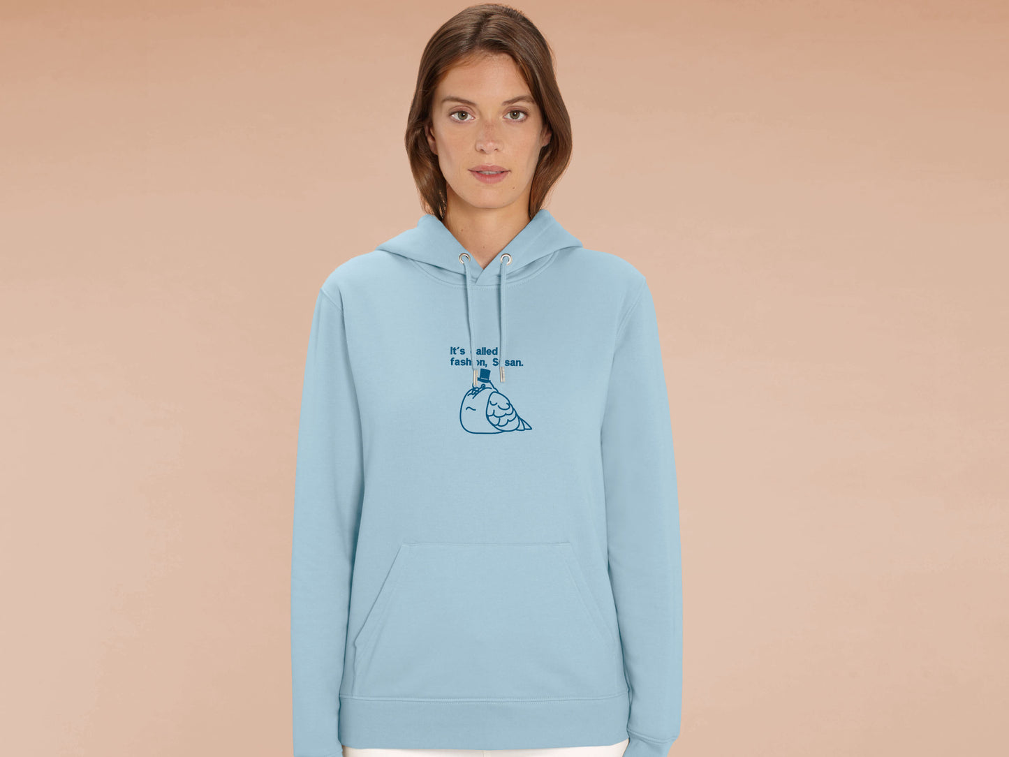A woman wearing a blue long sleeve fleece hoodie, with an embroidered blue thread design of cute fat pigeon wearing a top hat with text underneath reading It's Called Fashion, Susan.