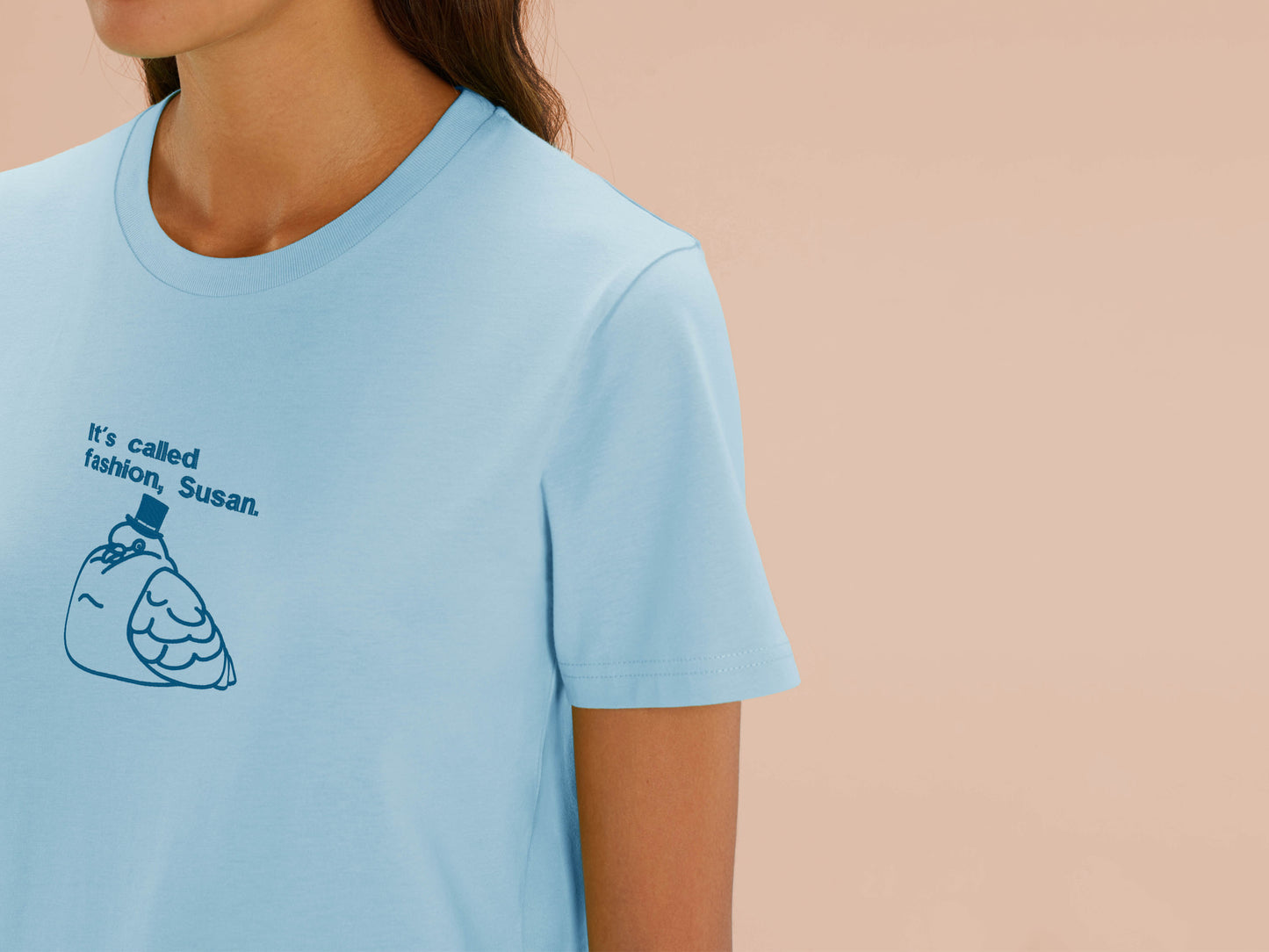 A woman wearing a blue crew neck short sleeve t-shirt, with an embroidered blue thread design of cute fat pigeon wearing a top hat with text underneath reading It's Called Fashion, Susan.