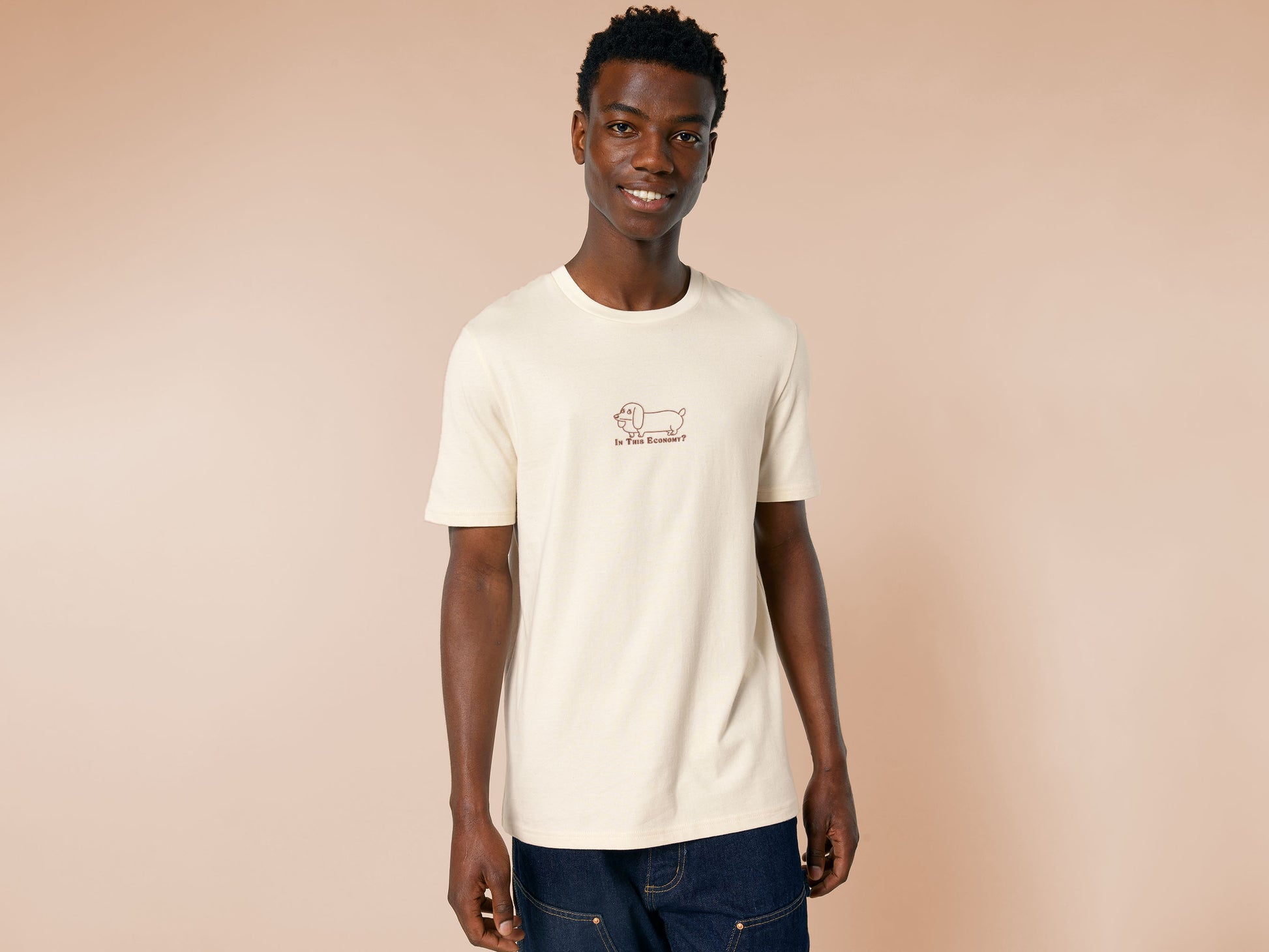 A cream crew neck short sleeve t-shirt, with an embroidered brown thread design of a cute daschund dog looking concerend surrounded by the quote In This Economy?