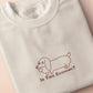 A cream crew neck short sleeve t-shirt, with an embroidered brown thread design of a cute daschund dog looking concerend surrounded by the quote In This Economy?