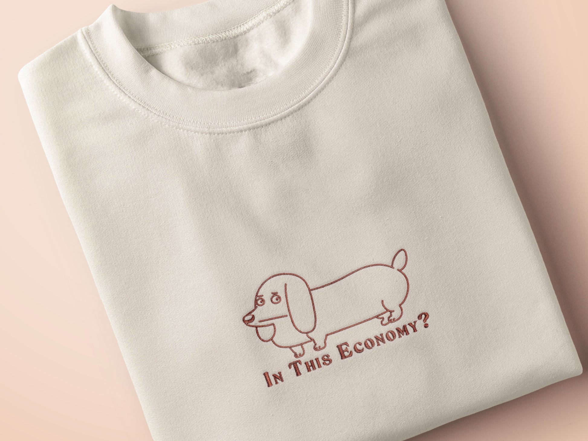 A cream crew neck short sleeve t-shirt, with an embroidered brown thread design of a cute daschund dog looking concerend surrounded by the quote In This Economy?