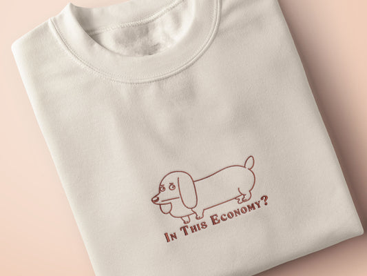 A cream crew neck short sleeve t-shirt, with an embroidered brown thread design of a cute daschund dog looking concerend surrounded by the quote In This Economy?