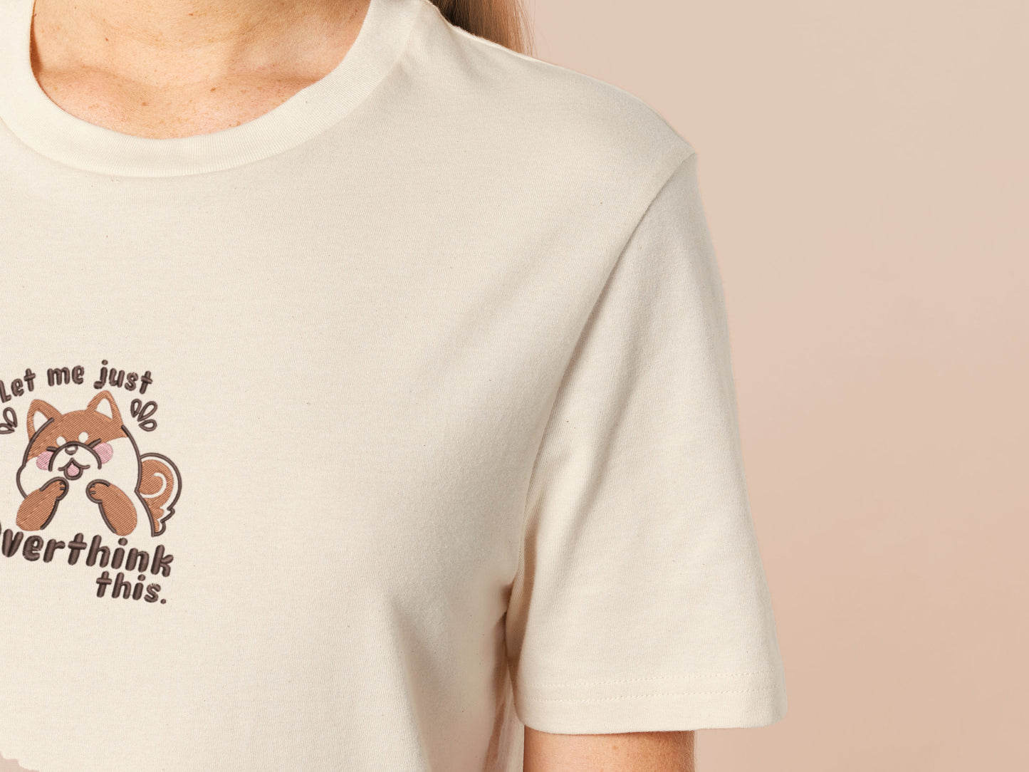 A cream crew neck short sleeve t-shirt, with an embroidered design of a cute shiba inu dog looking stressed surrounded by the quote Let Me Just Overthink This