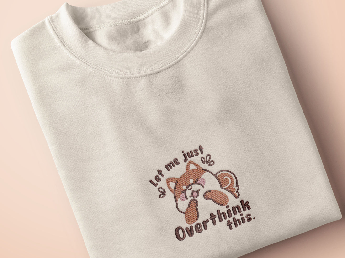 A cream crew neck short sleeve t-shirt, with an embroidered design of a cute shiba inu dog looking stressed surrounded by the quote Let Me Just Overthink This