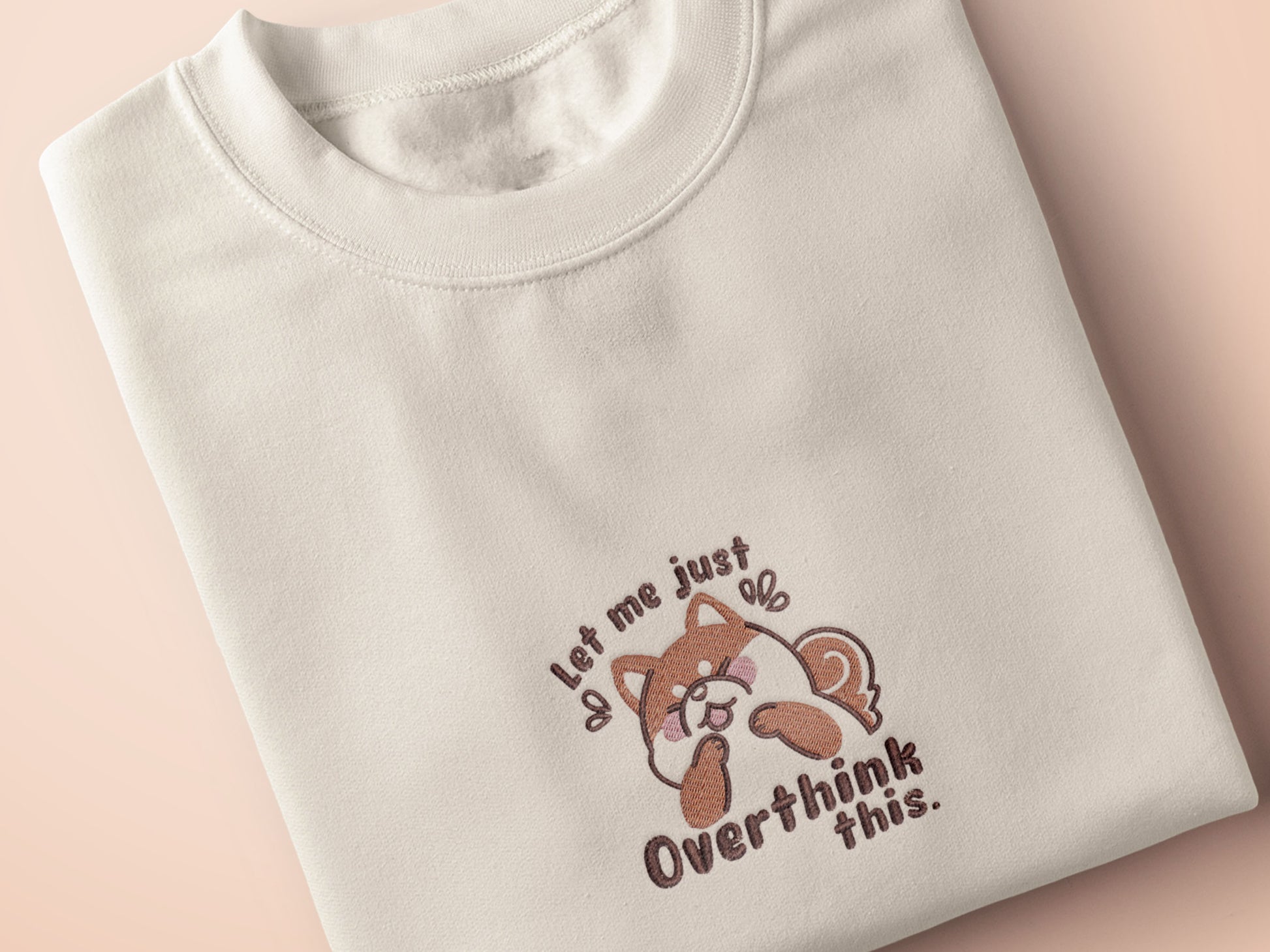 A cream crew neck short sleeve t-shirt, with an embroidered design of a cute shiba inu dog looking stressed surrounded by the quote Let Me Just Overthink This