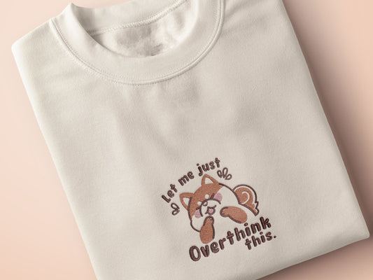 A cream crew neck short sleeve t-shirt, with an embroidered design of a cute shiba inu dog looking stressed surrounded by the quote Let Me Just Overthink This