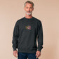 A dark grey crew neck long sleeve sweater, with an embroidered cute chibi mimic chest holding a D20 dice rolled a one and the quote Looks fine to you.