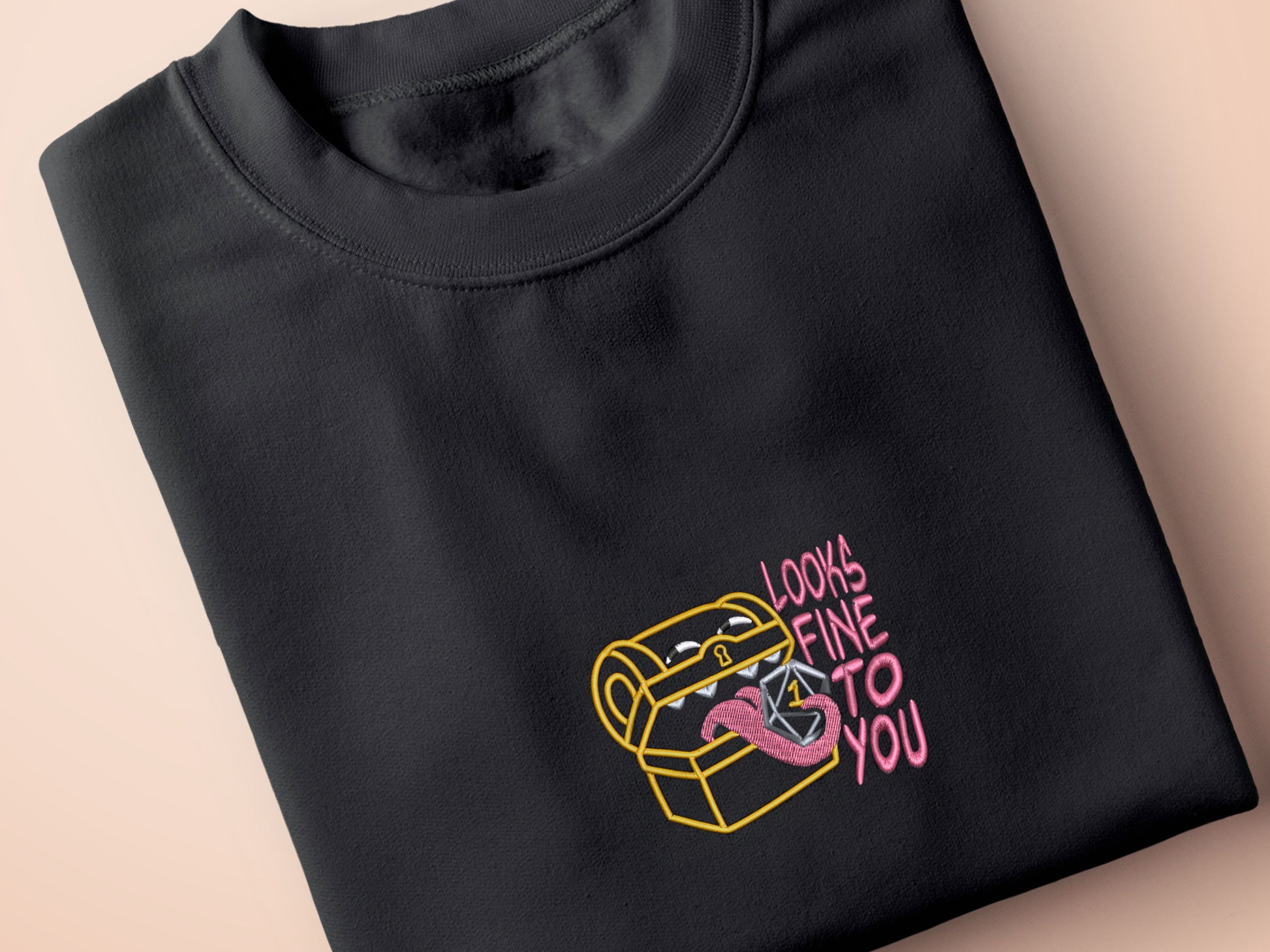 A dark grey crew neck short sleeve t-shirt, with an embroidered cute chibi mimic chest holding a D20 dice rolled a one and the quote Looks fine to you.