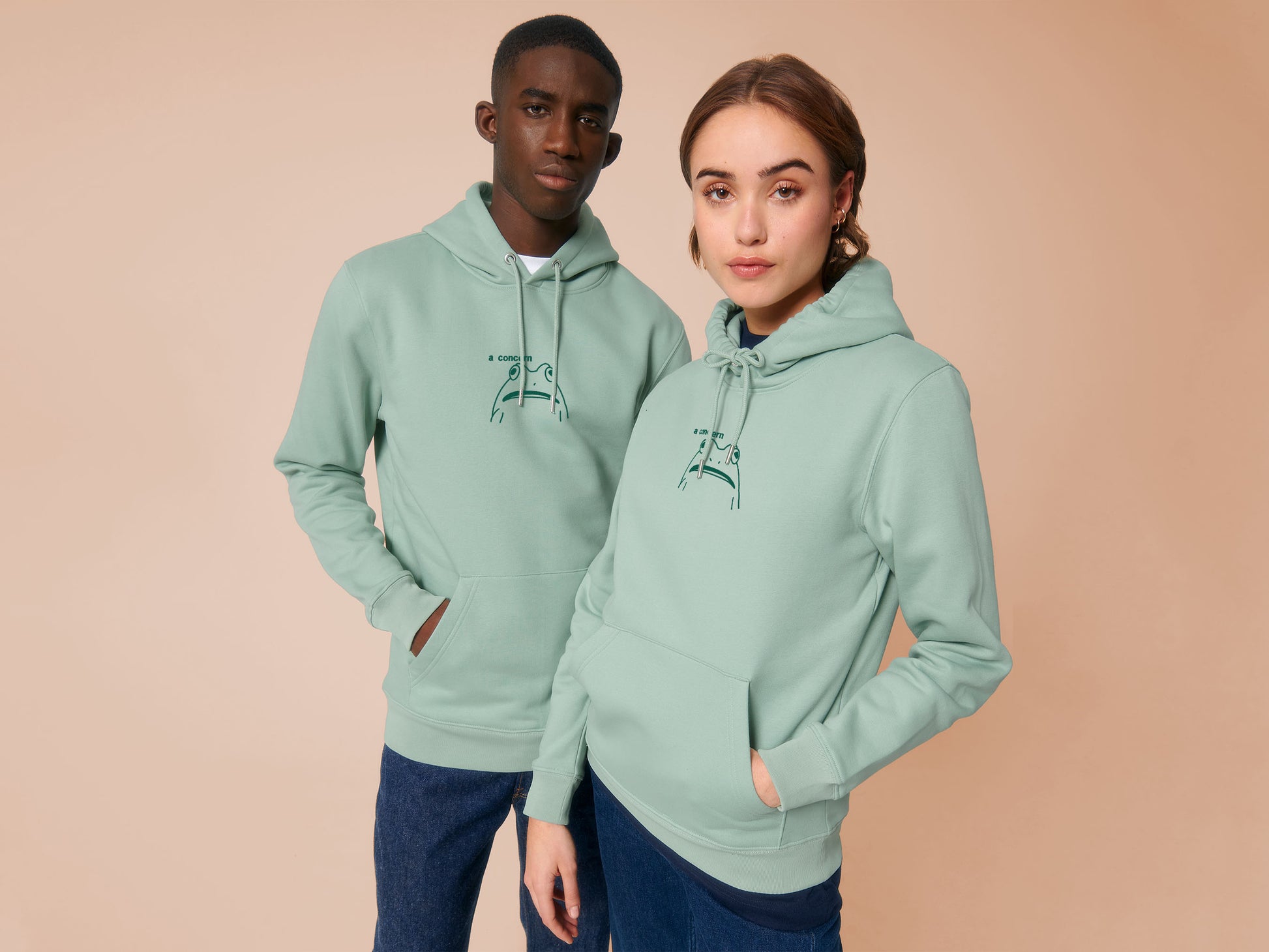 A woman and man both wearing an green long sleeve fleece hoodie, with an embroidered black thread design of cute confused looking frog with the text a concern.