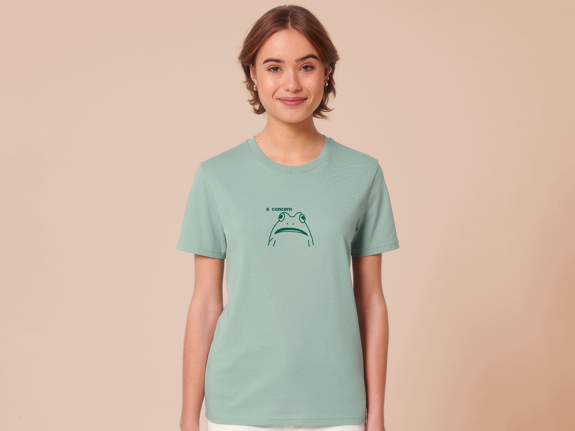 A woman wearing a green crew neck short sleeve t-shirt, with an embroidered green thread design of cute confused looking frog with the text a concern.