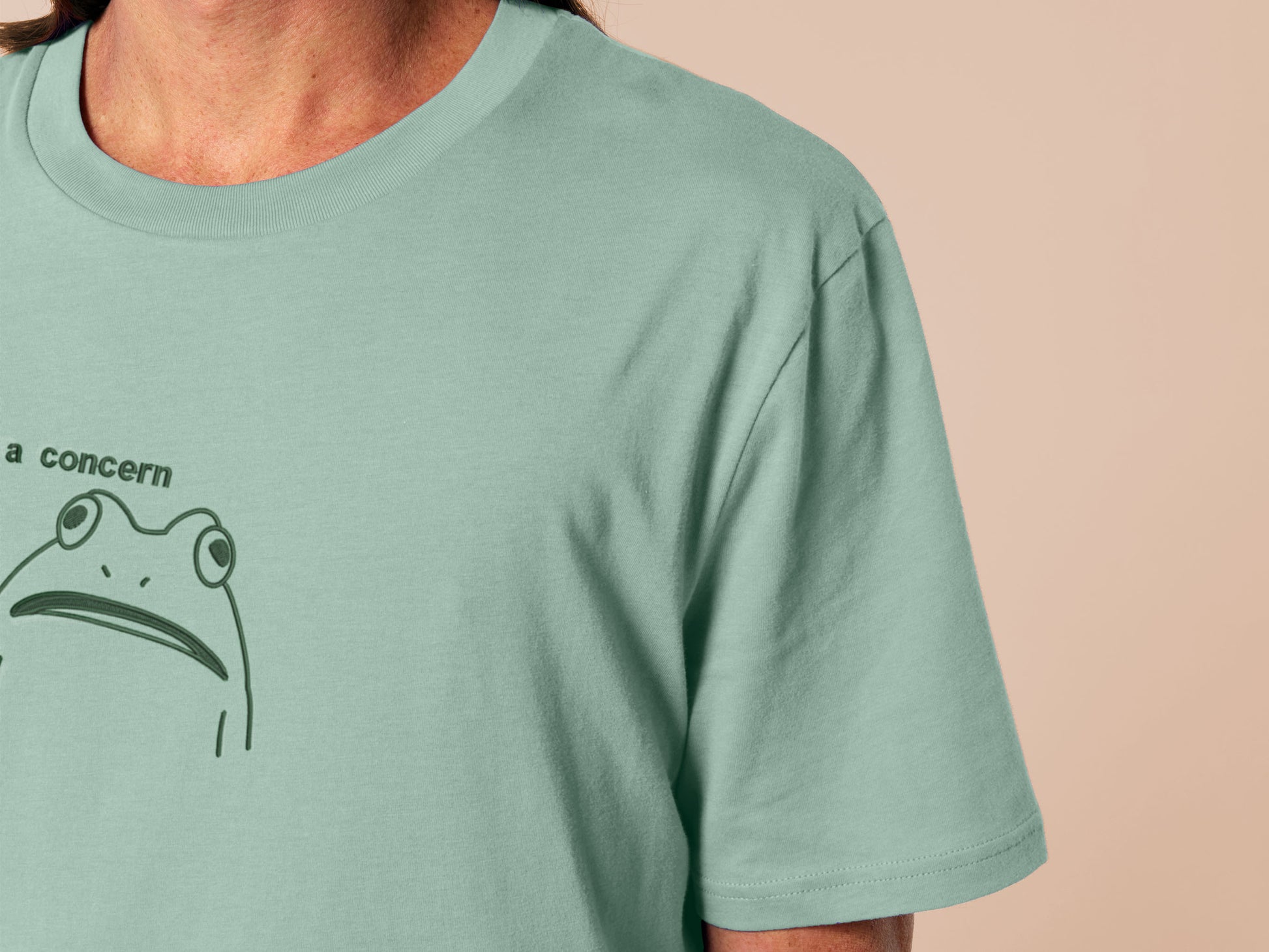 An embroidered green t-shirt design of a funny cartoon frog with the text a concern.