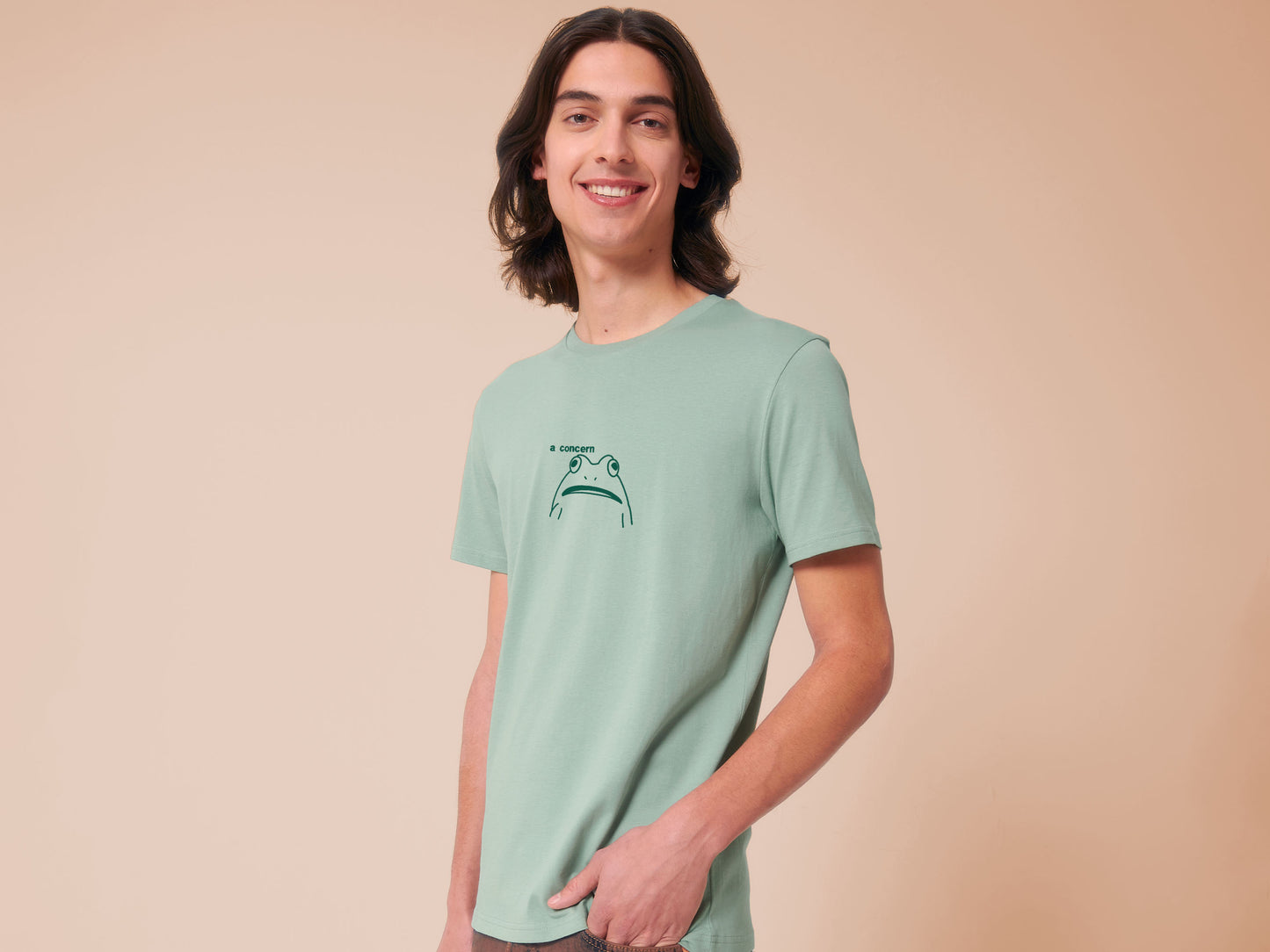 A man wearing a green crew neck short sleeve t-shirt, with an embroidered green thread design of cute confused looking frog with the text a concern.